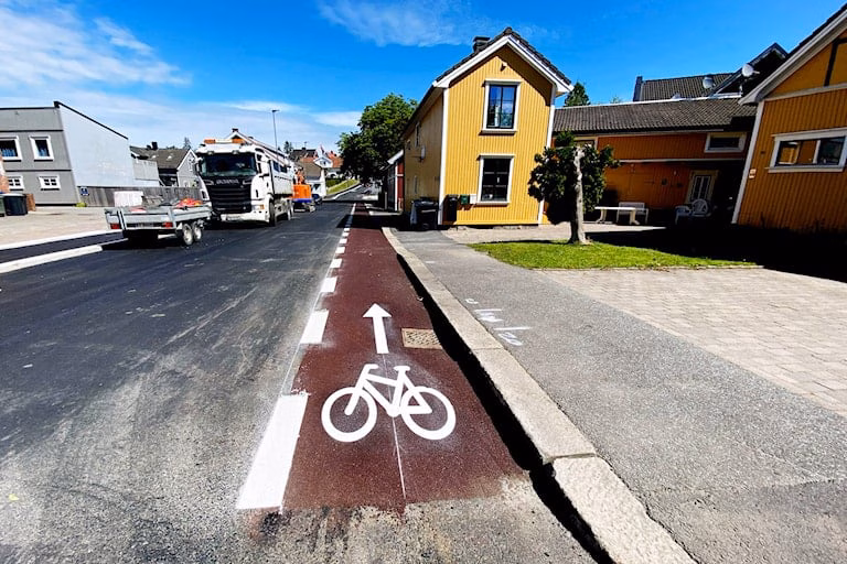 Rødt sykkelfelt midt i bildet med bilveg til høyre og så et fortau. Til høyre for det røde sykkelfeltet er det en gårdsplass og et gult hus. Bebyggelse langs vegen. 